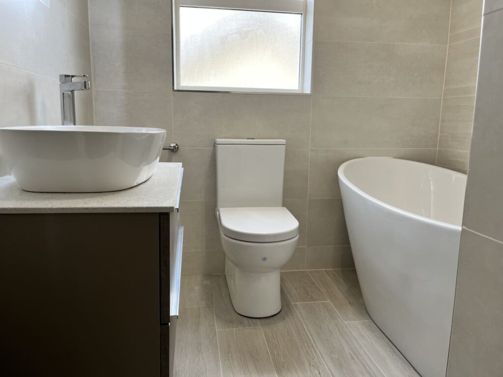 Modern bathroom featuring a freestanding tub, wall-mounted sink, and sleek toilet against light tiled walls. Pro Renovations 