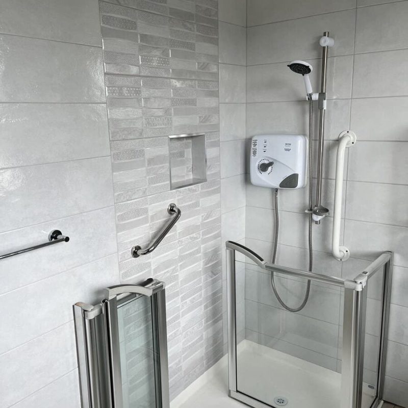 Accessible shower with glass door, grab bars, and a handheld showerhead, featuring light-colored tiled walls in the Gallery