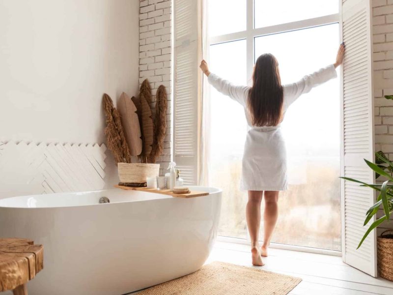 A woman in a white robe opens shutters in a bright bathroom with a freestanding tub and greenery, ideal for family bathroom r