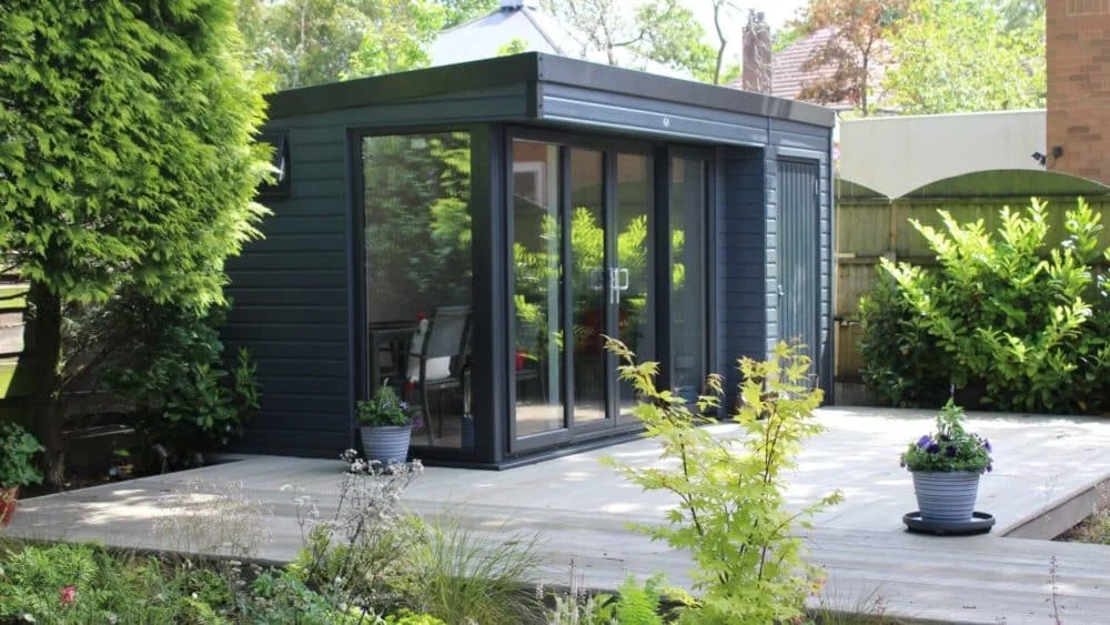 Lush garden with manicured shrubs, a modern shed, and two people seated on a patio surrounded by greenery and flowers